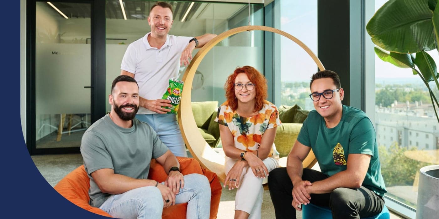 Four people in an office space during the daytime. The person on the far left is sitting in an orange bean bag chair; they wear a green shirt and light-blue jeans. The person standing directly behind them wears a polo shirt and jeans; they hold a bag of chips. The person in the middle sits in a circular chair; they have orange hair and wear a white shirt with a floral pattern and white jeans. The person to the far right sits on a blue pouff; they wear a green shirt, black jeans, and glasses.