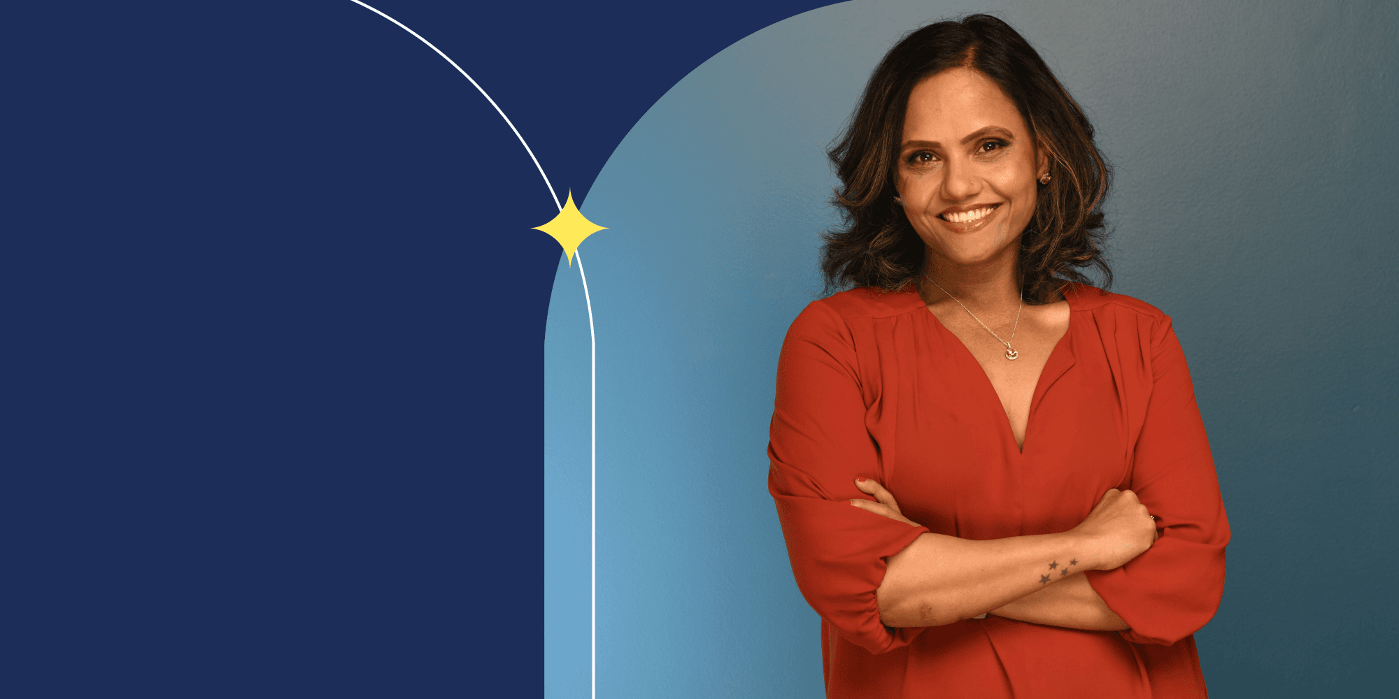 A person with their arms crossed and smiling at the camera. They have wavy brown hair; they're wearing an orange shirt and a silver necklace.